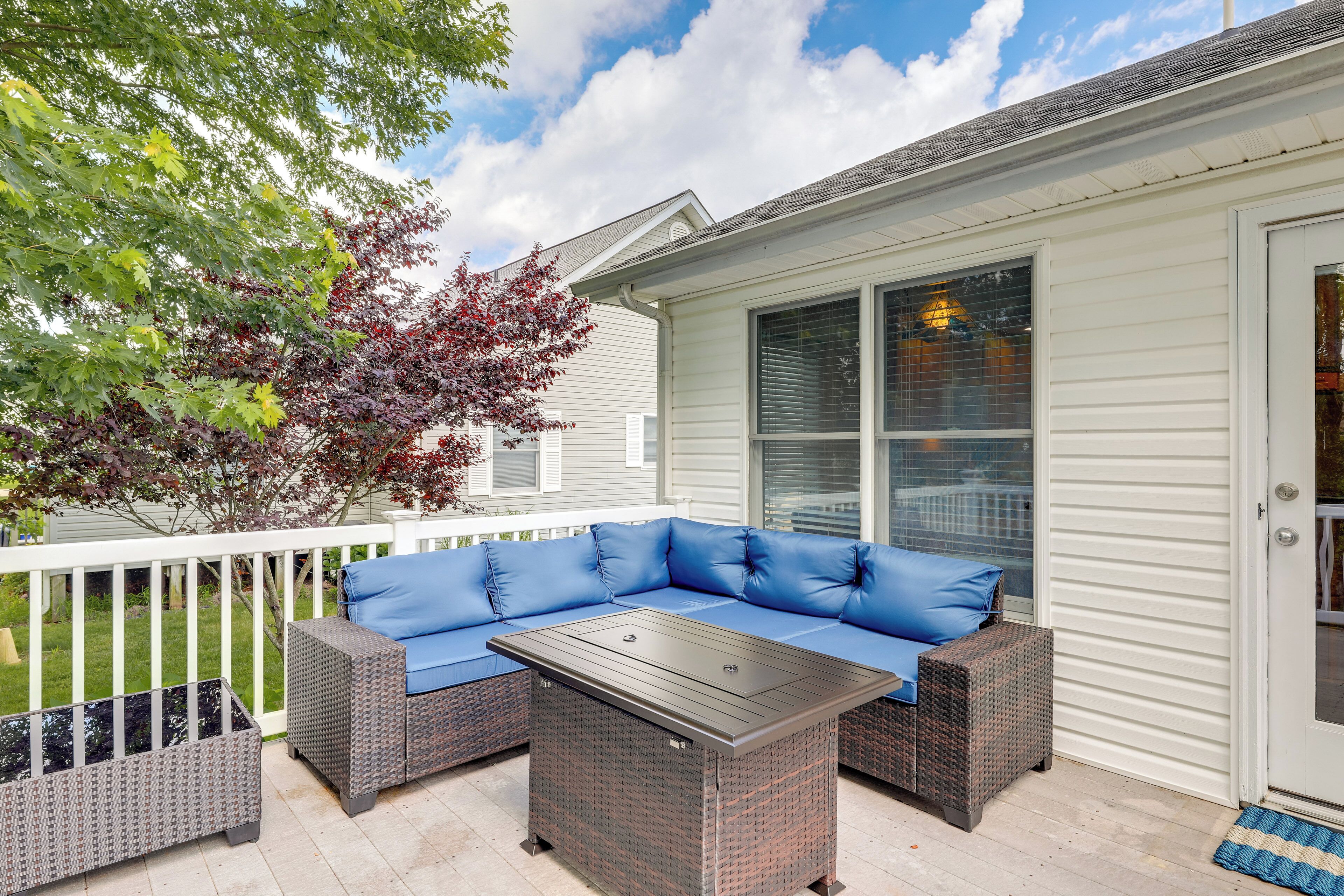 A covered patio with a sectional sofa and a table, surrounded by a white railing and a tree with red leaves.