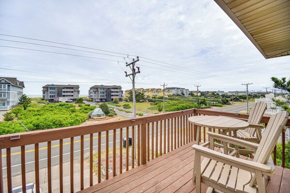 A wooden deck with a table and chairs, overlooking a road with buildings and greenery. - Walk to Beach + Restaurants: Outer Banks Retreat (Kill Devil Hills)