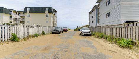 A sandy road flanked by two-story buildings with balconies, parked cars, and a wooden fence.
