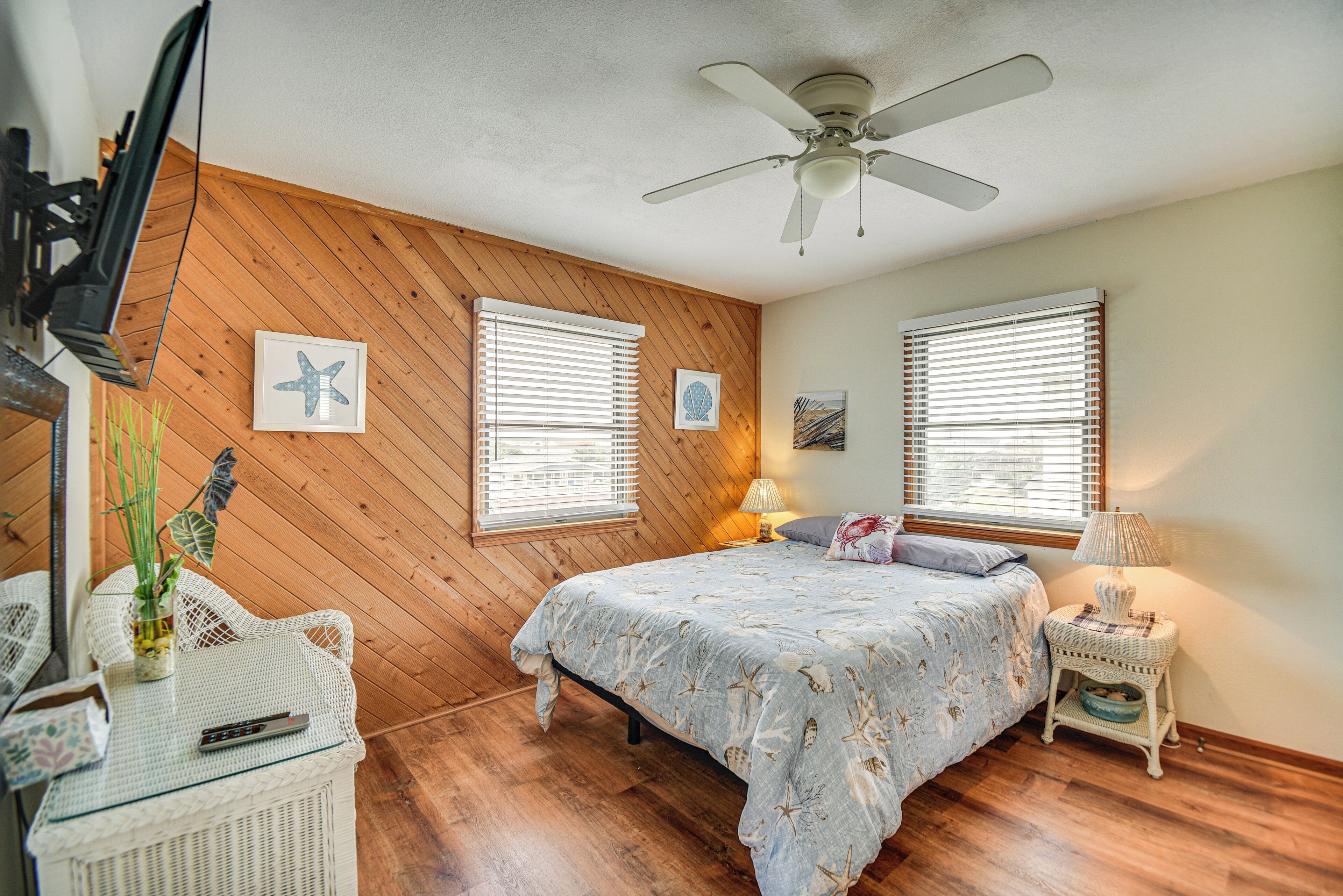 A bedroom with a wooden wall, two windows with blinds, a bed with a floral bedspread, a ceiling fan, and a small table with a lamp.