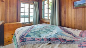 Dormitorio de cabaña de madera con cama, cómoda de madera y ventana con vista a un cuerpo de agua.