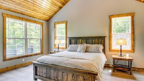 A bedroom with a wooden bed, two bedside tables with lamps, and large windows with wooden frames. - 11 Mi to State Park: Home w/ Dock in Mill Spring! (Mill Spring)