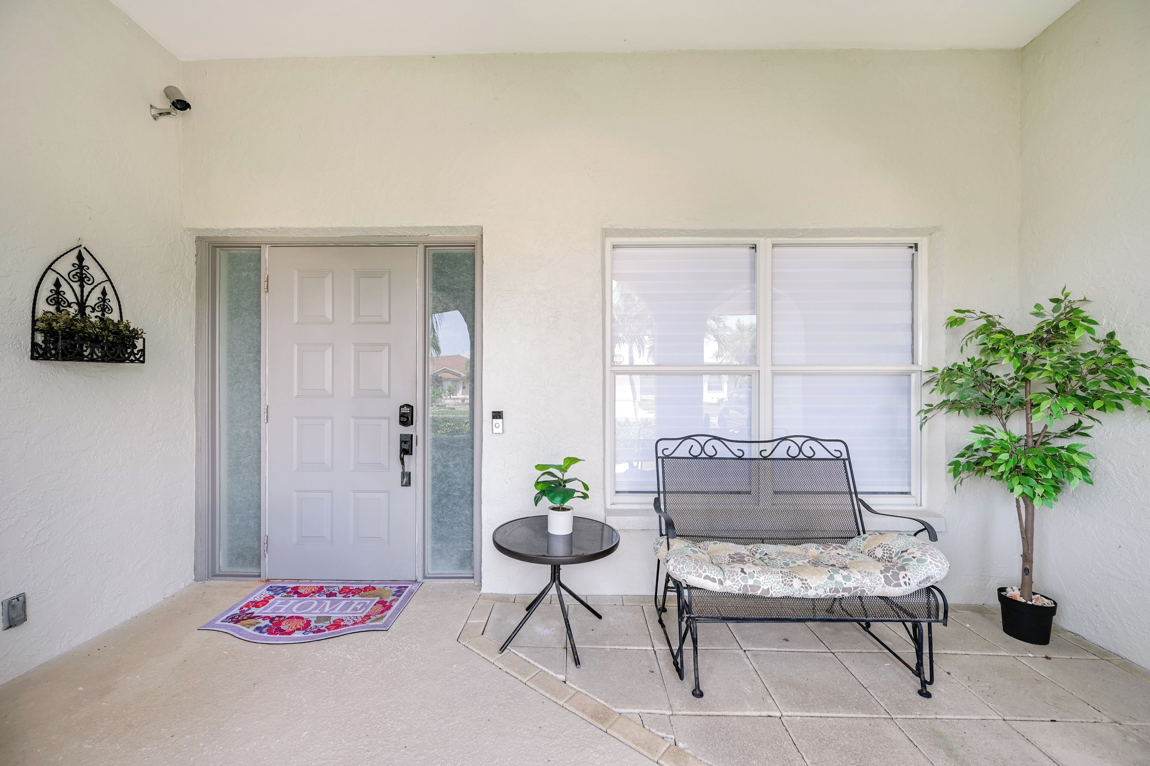 A clean, well-maintained entrance with a white door, a small round table with a potted plant, a metal chair, and a potted tree.