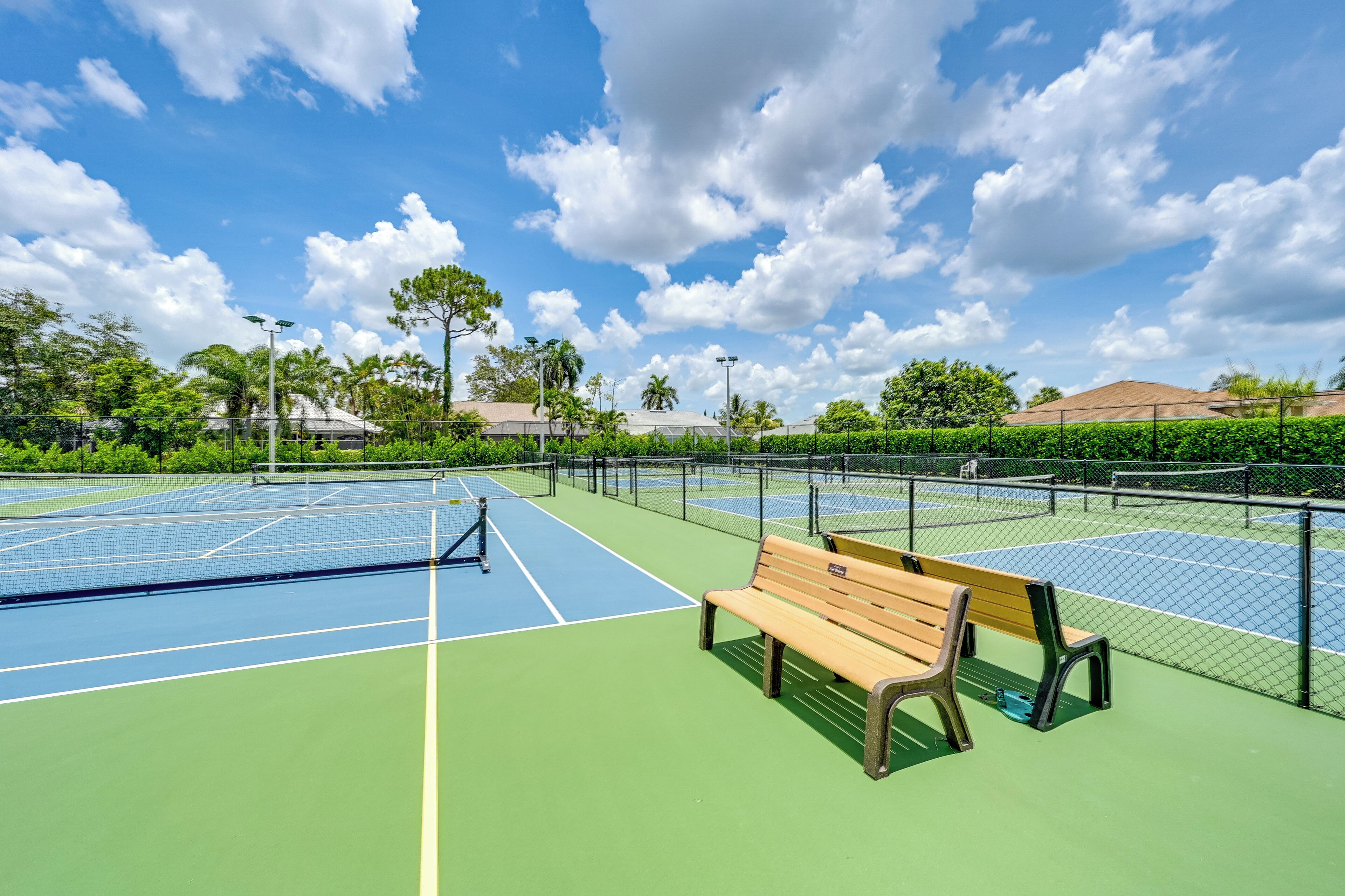 Un court de tennis avec un banc et une clôture, entouré de verdure et de bâtiments.
