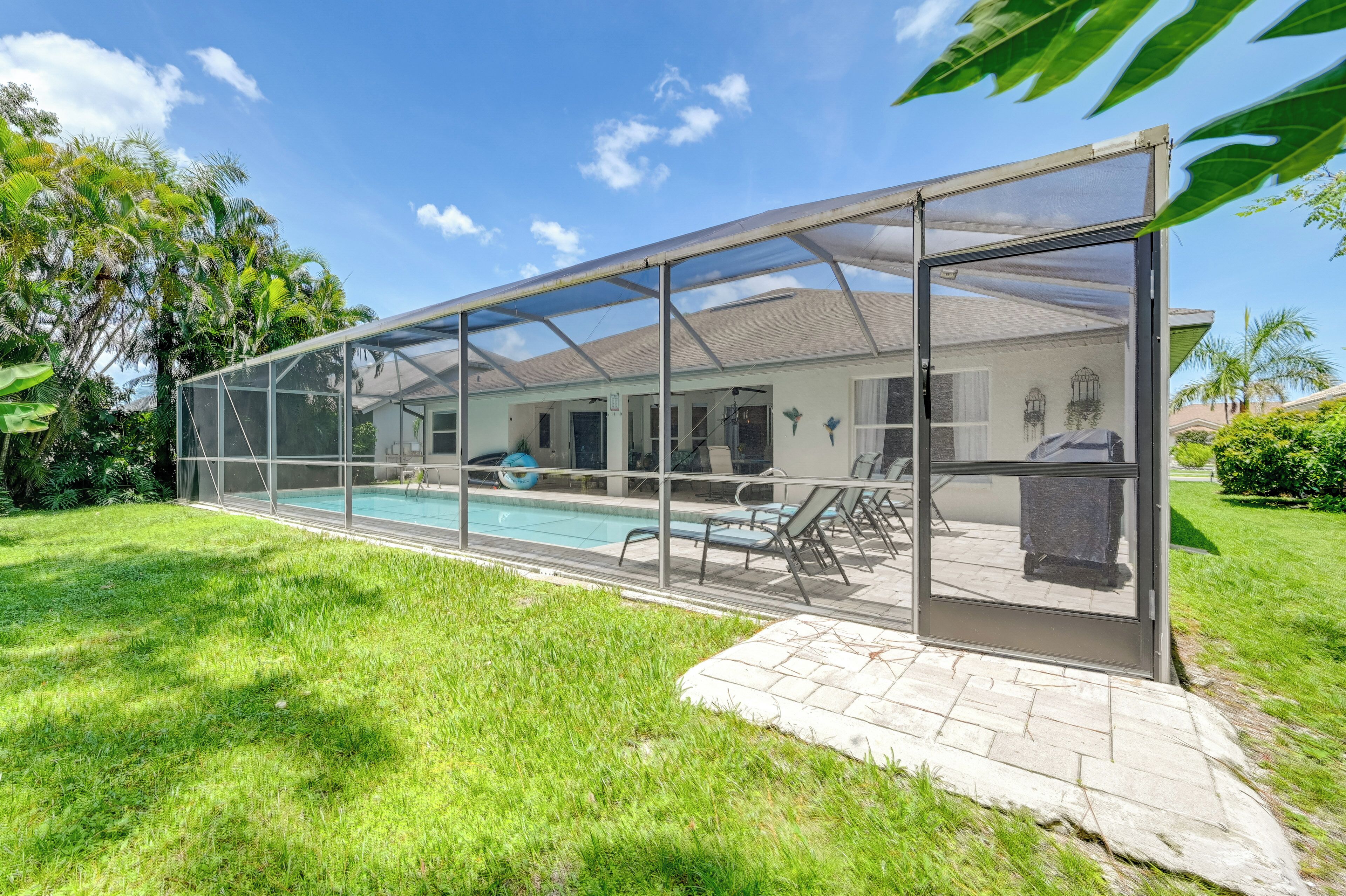 A covered swimming pool with a glass enclosure, a patio area with chairs, and a view of a house in the background.