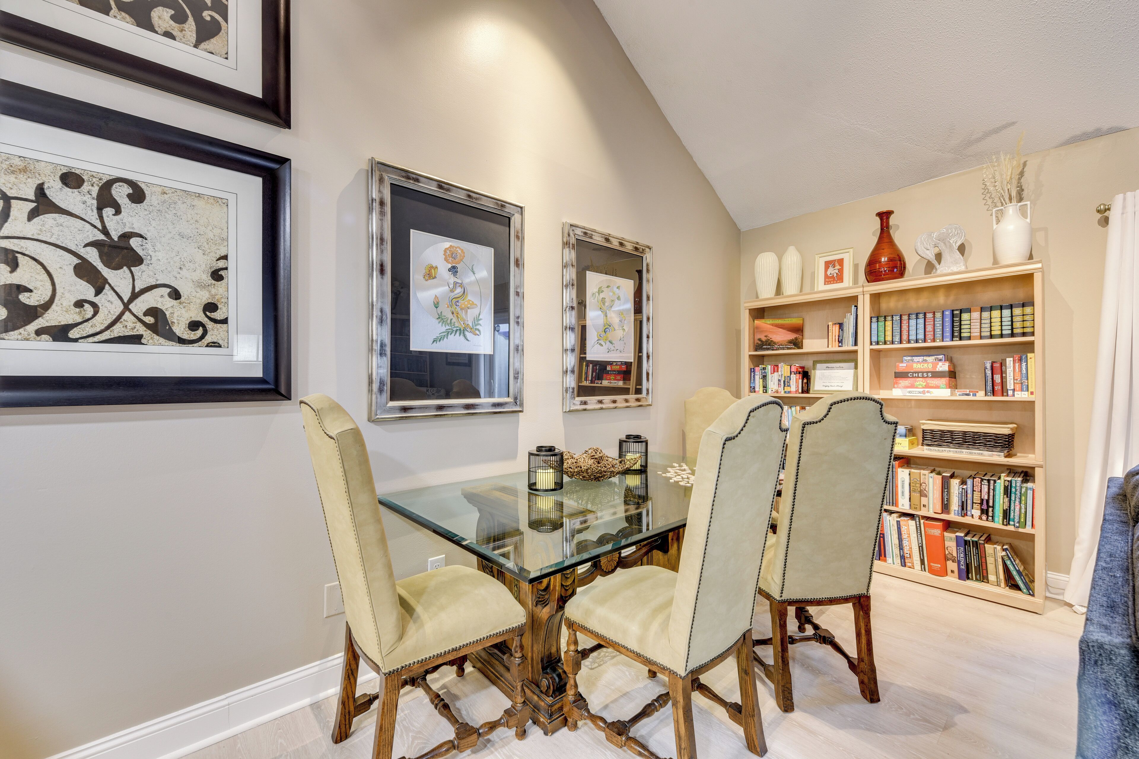 Un coin repas avec une table en verre et quatre chaises, une bibliothèque remplie de livres et des œuvres d’art encadrées accrochées au mur.