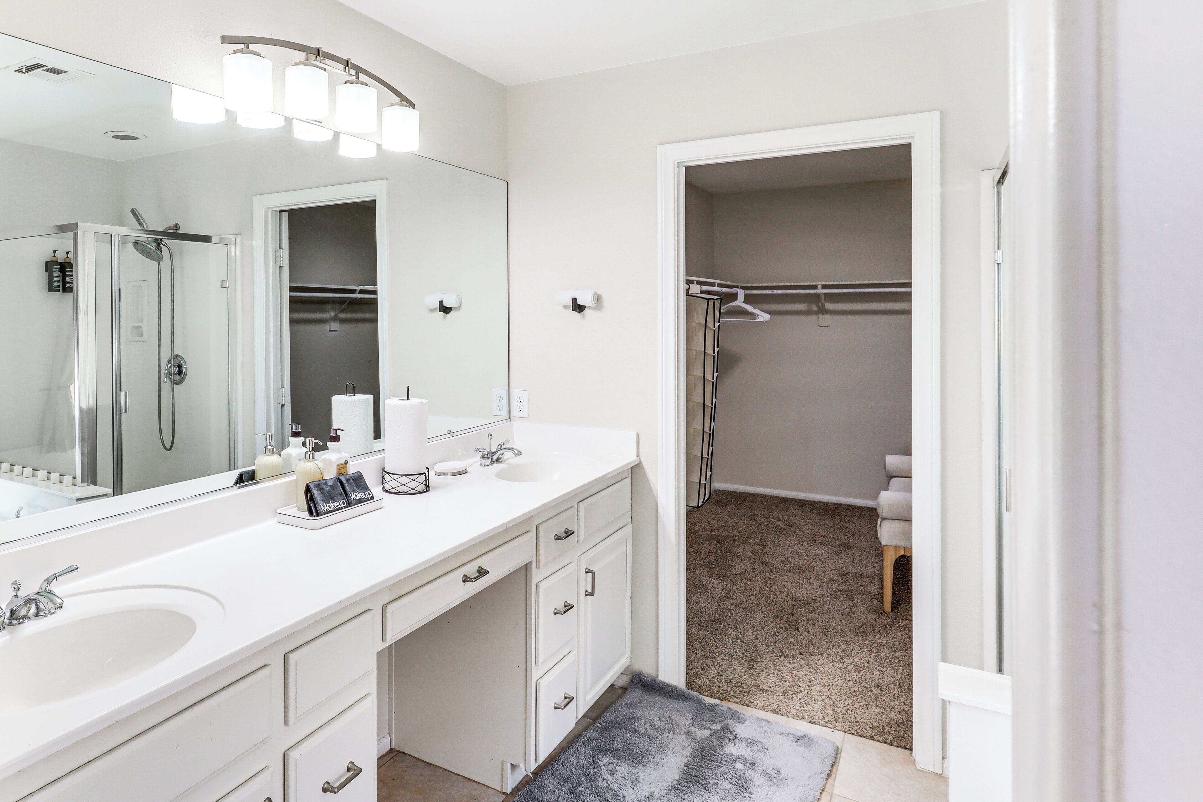 A modern bathroom with a large mirror, two sinks, a shower, and a walk-in closet.
