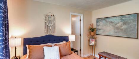 A bedroom with a large bed, bedside lamps, a headboard, a bench, a wall-mounted artwork, and a door leading to another room.