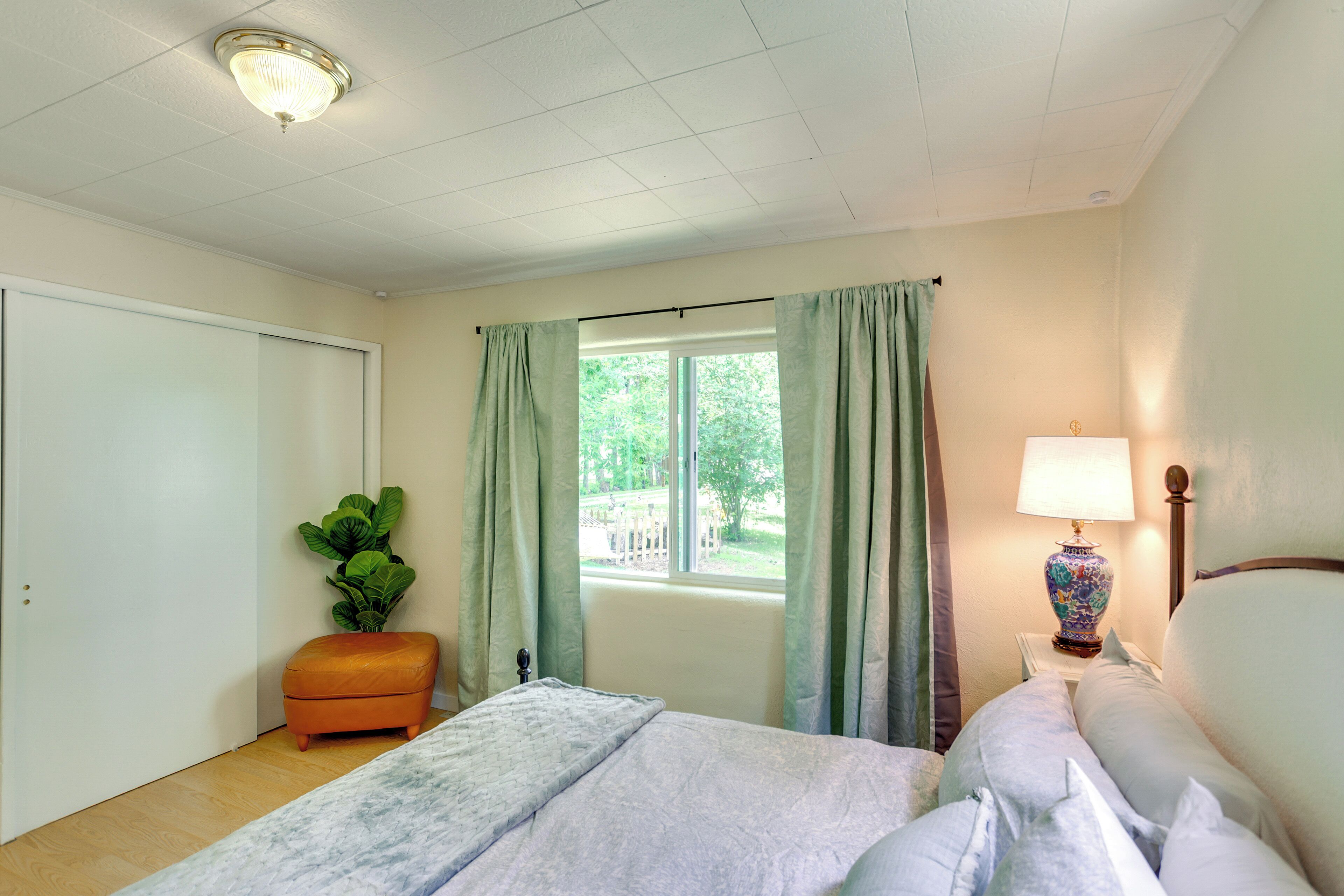 Ein Schlafzimmer mit einem Bett, einem Nachttisch mit Lampe, einem Fenster mit Vorhängen, einer Pflanze und einer Tür.