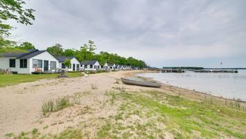 A row of white cabins with green trim, a sandy beach, and a calm lake with a pier in the background.