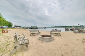 Ein Strand mit Holzbanken und einer Feuerstelle, mit Blick auf einen ruhigen See mit Anlegestellen und Booten.