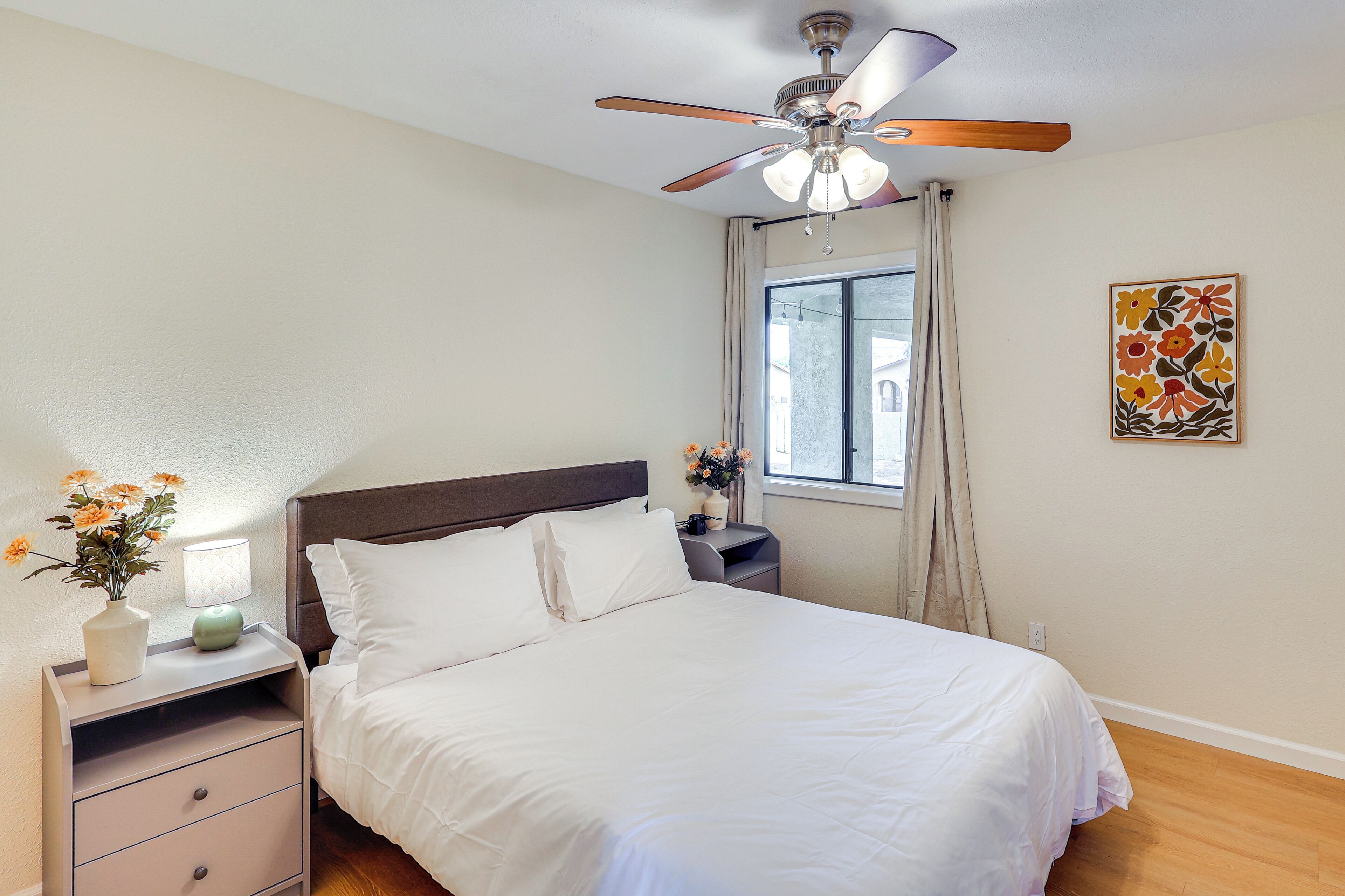 A neatly made bed with white linens, a wooden headboard, a nightstand with a lamp, and a ceiling fan.