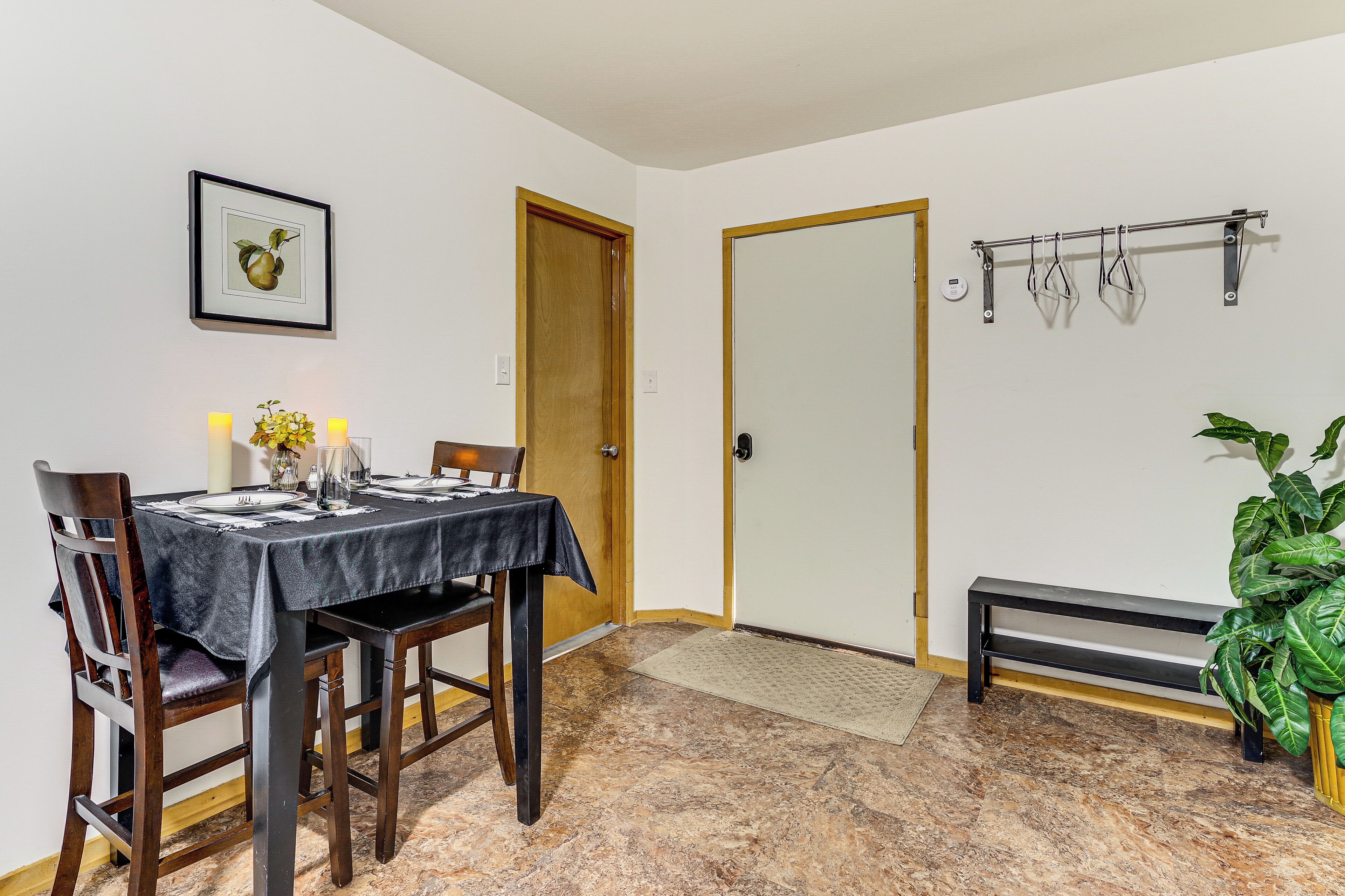 Un coin repas avec une table dressée pour quatre personnes, une porte en bois, un banc, une étagère fixée au mur et une plante en pot.