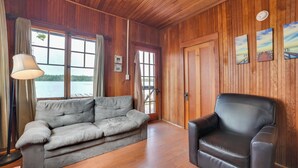 Una habitación con paneles de madera, un sofá gris y un sillón negro, ventanas amplias con cortinas, y vista a un cuerpo de agua.