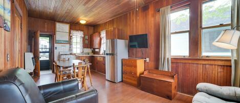 A cozy cabin interior with a wooden ceiling, a dining area, a kitchen with a refrigerator and TV, and a seating area with a sofa and a chair.