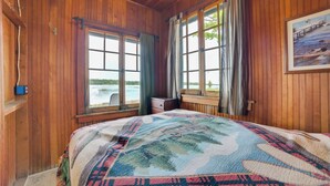 Una habitación de cabaña de madera con una cama grande, dos ventanas con cortinas, un cuadro en la pared y vistas a un lago.