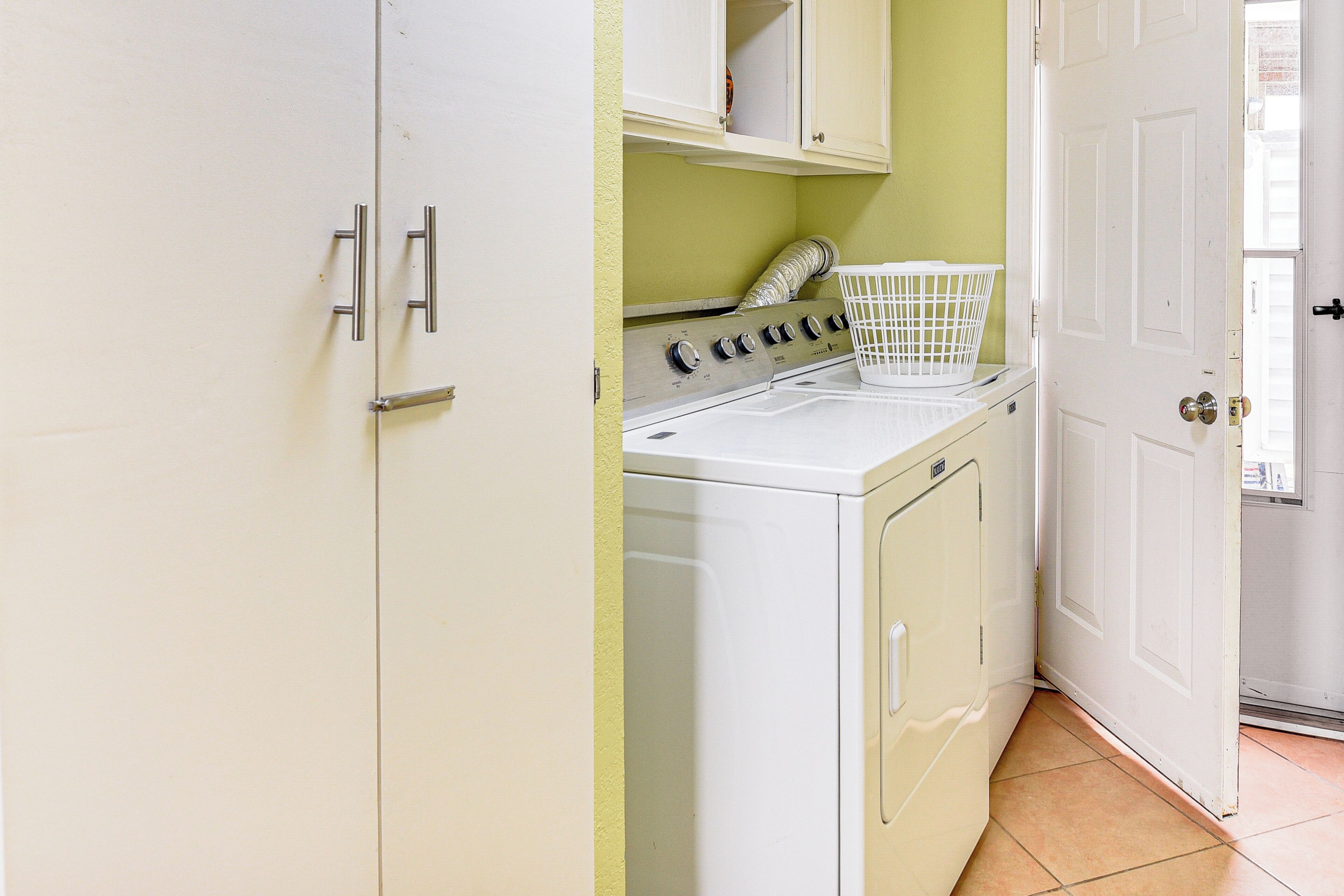 A laundry room with a white washing machine and dryer, a white basket, and a door leading to another room.