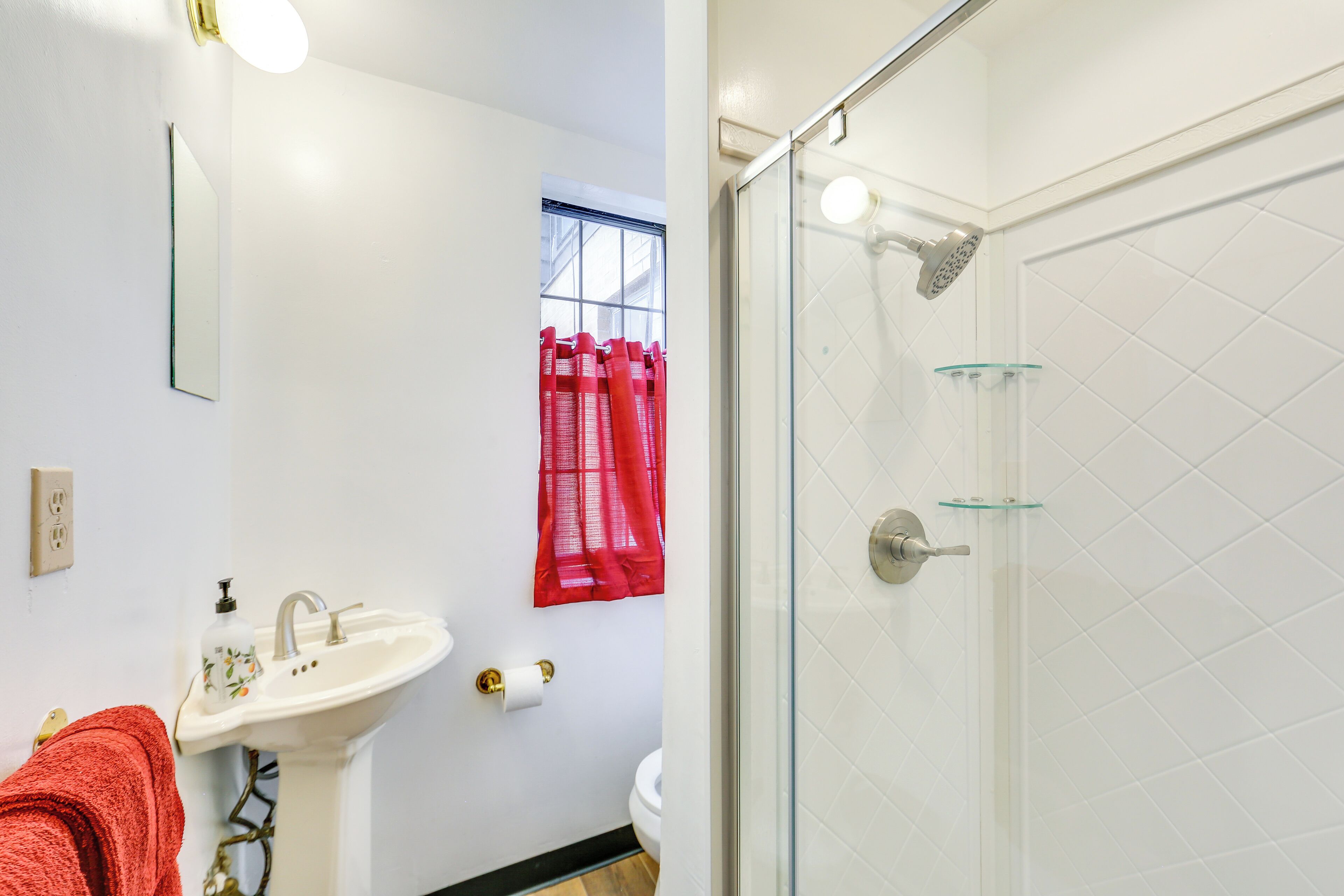 Une salle de bain avec un lavabo blanc, un miroir, une serviette rouge, une cabine de douche en verre et une fenêtre avec des rideaux rouges.