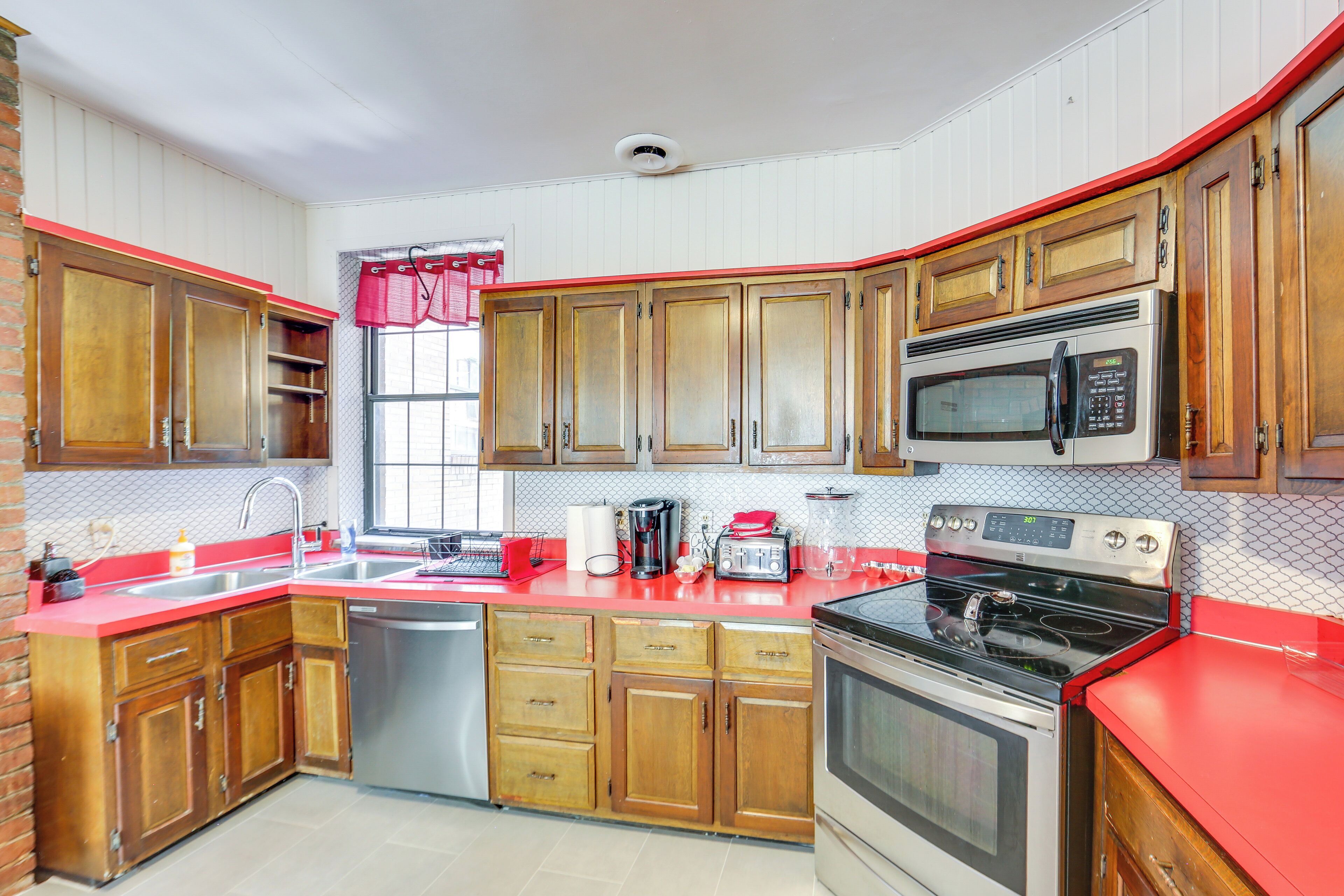 A kitchen with wooden cabinets, a red countertop, stainless steel appliances, and a window with a red curtain.