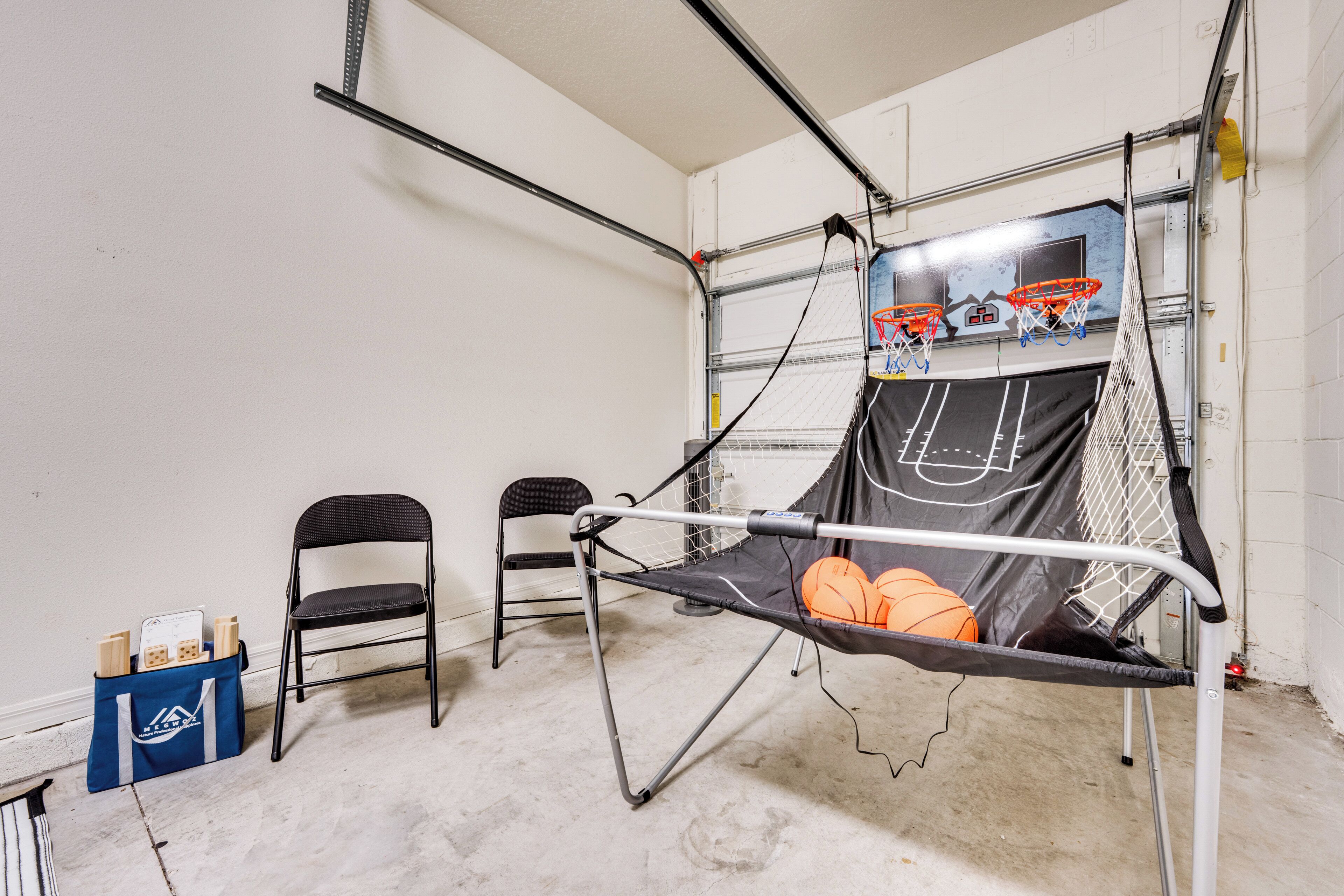 Un panier de basketball est installé dans une pièce aux murs blancs et au sol en béton. On y trouve deux chaises et un sac bleu contenant divers objets, posés sur le sol.