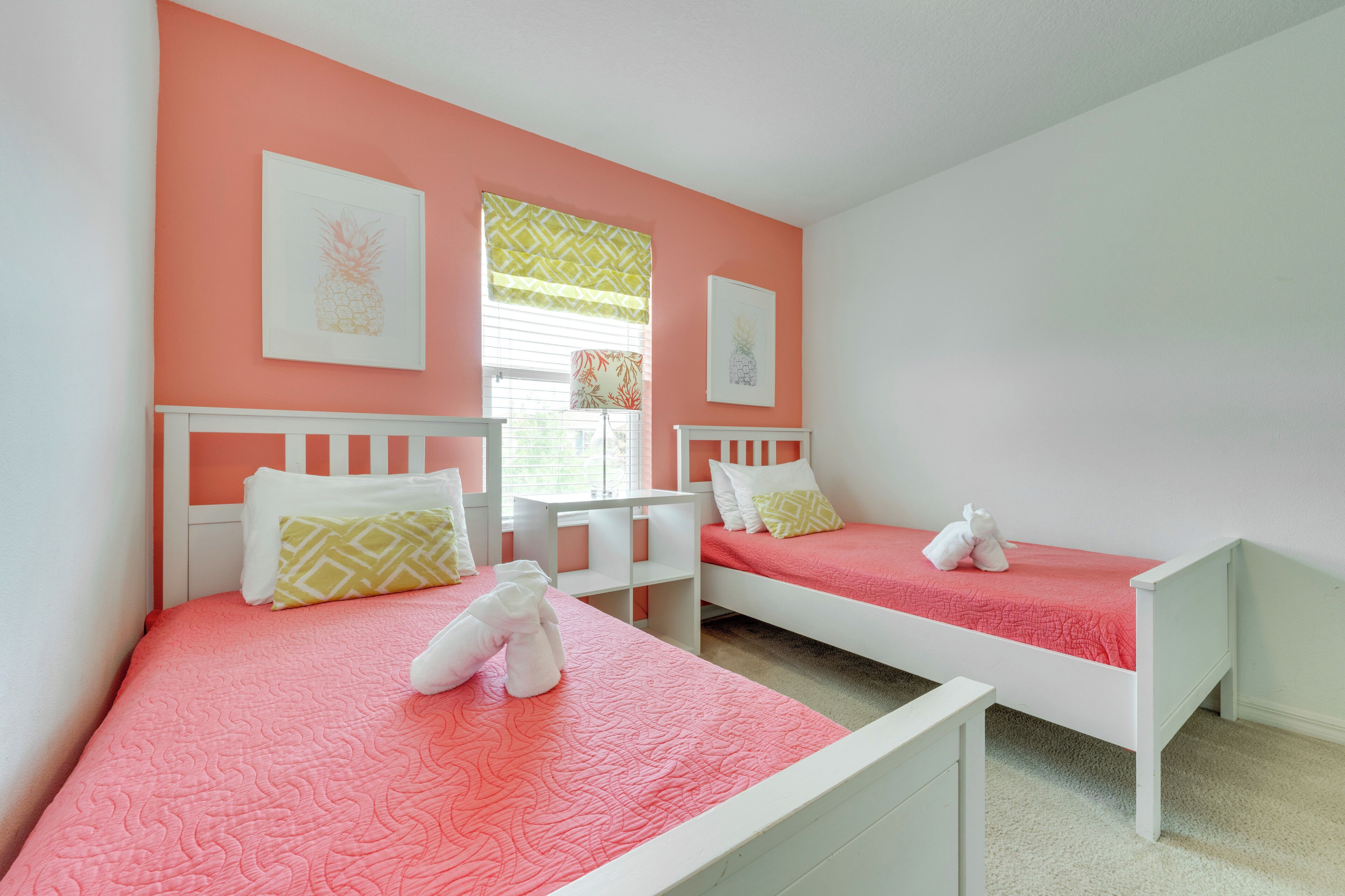 A room with two white beds, each with a pink bedspread and pillows. There are framed pictures on the walls, and a window with blinds.
