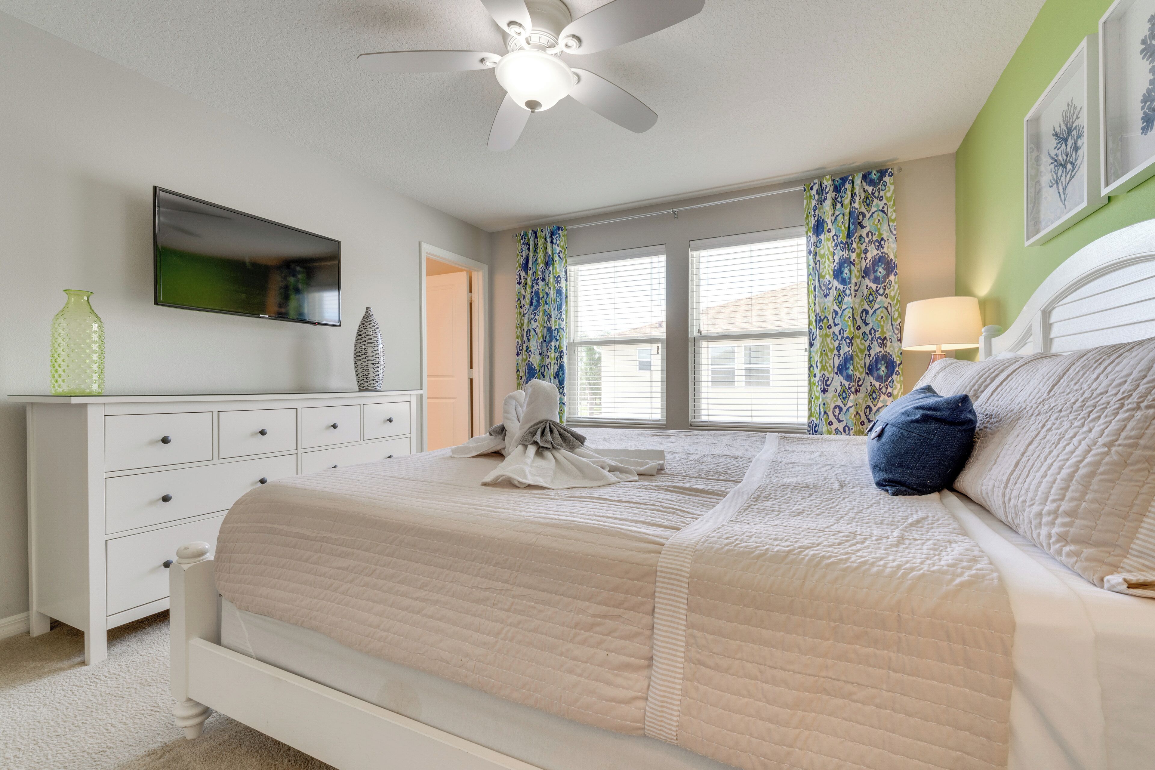 A bedroom with a bed, a ceiling fan, a TV, a dresser, and a window with curtains.