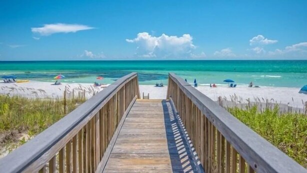 A wooden boardwalk leading to a beach with clear turquoise water and a few umbrellas.