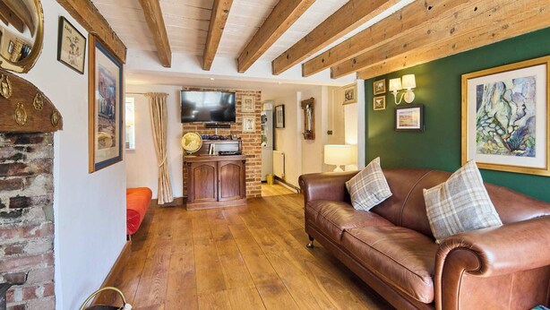 A cozy living room with a leather sofa, wooden beams, a brick fireplace, and a television. - Host Stay Romney Cottage (Norwich)