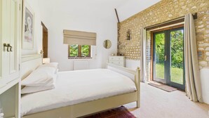 A bedroom with a bed, a dresser, a window with a view of greenery, a stone wall, and a door leading outside. - Host Stay The Old Foundry (King's Lynn)