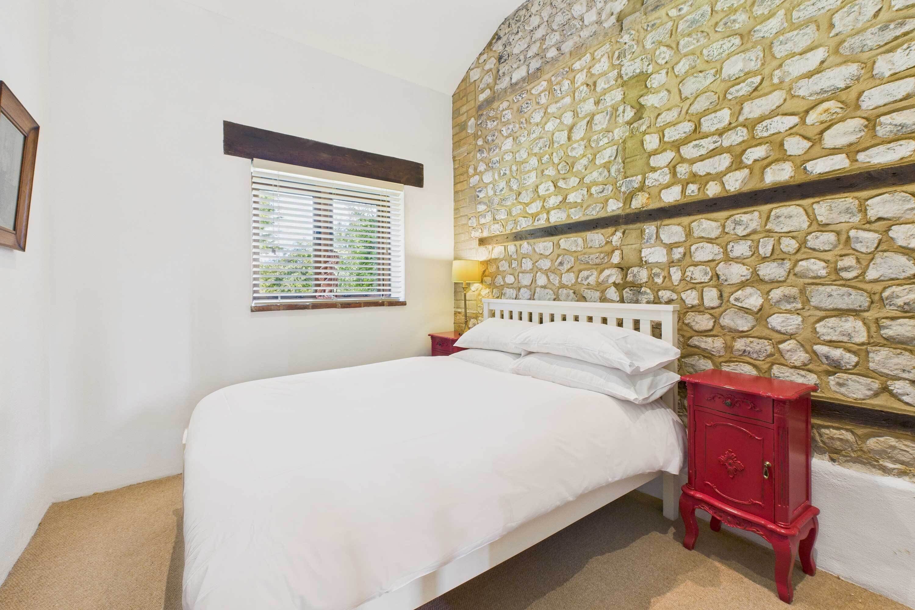 A bedroom with a stone wall, a bed with white bedding, a red nightstand, and a window with blinds.