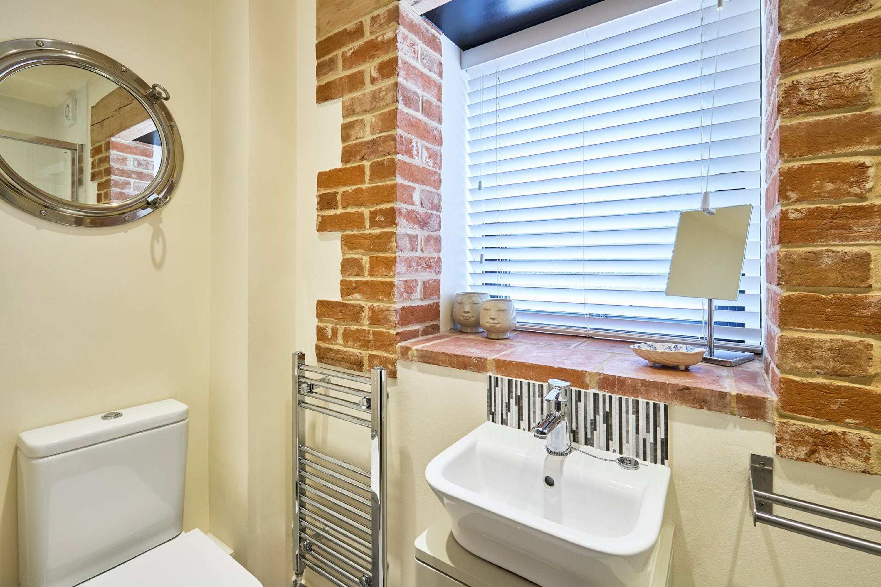 A bathroom with a white sink, a round mirror, and a window with blinds.
