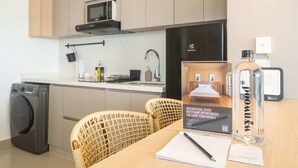 Cocina moderna equipada con lavadora, microondas y refrigerador. Se observa una silla de mimbre y una mesa con libreta, bolígrafo y una botella de agua.