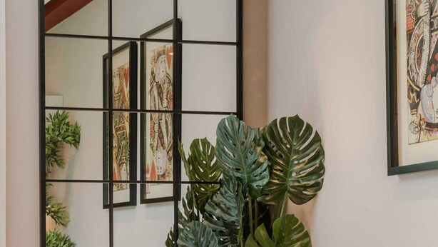 A living room with a sofa, decorative pillows, a side table with a lamp, and a large mirror with artwork. - The Honourable - Bloomsbury Square By Frankie Says (London)