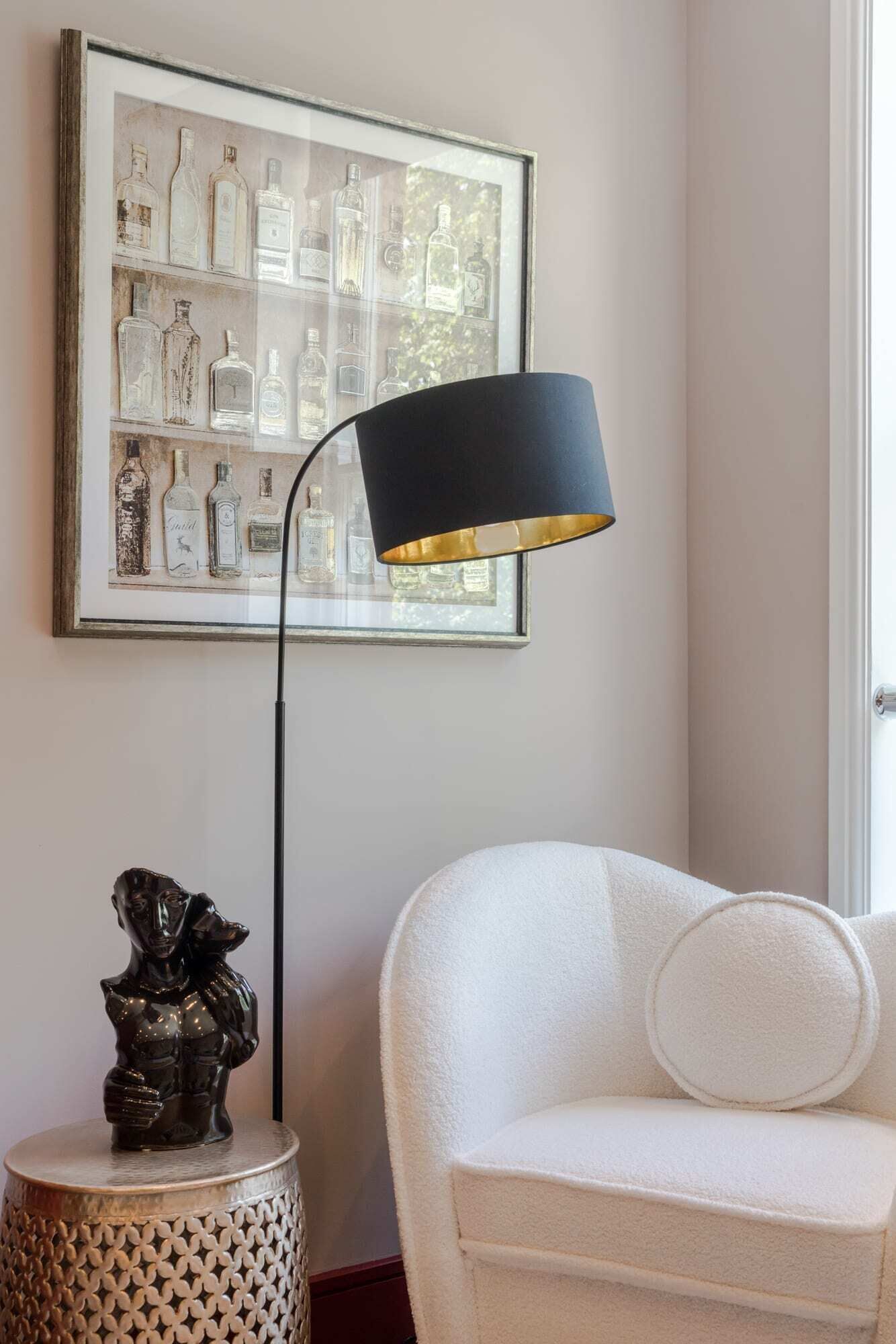 A framed artwork of bottles on the wall, a black floor lamp, a white sofa, and a small decorative sculpture on a round table.