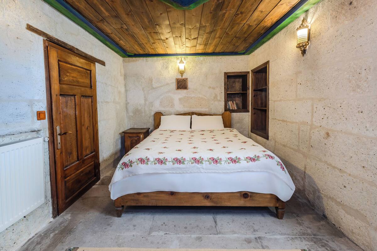 Ein Schlafzimmer mit einem hölzernen Bett, einem Nachttisch, einem Holzschrank, einem Heizkörper, einem an der Wand befestigten Regal mit Büchern und einer Holzbalkendecke.