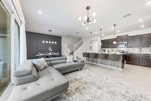 Una sala moderna con un sofá seccional gris, un comedor con barra y una cocina con armarios oscuros y un microondas.