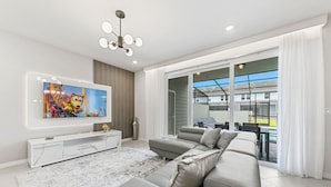 A modern living room with a grey sofa, a flat-screen TV mounted on the wall, a white entertainment unit, and a large sliding glass door leading to an outdoor area.