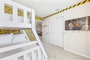 Een kinderbed met stapelbed en een Minion-thema matrasbeschermer, witte muren, een deur met een Minion poster en een gestreept plafond.