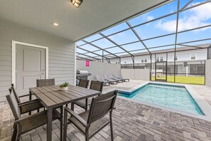 An outdoor patio with a pool, a table with chairs, and a grill area.