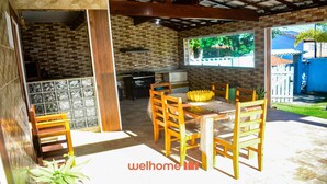 Un patio exterior con pared de ladrillo, sillas de madera, una mesa con centro de mesa de plátanos y vista a una casa azul a través de una ventana.