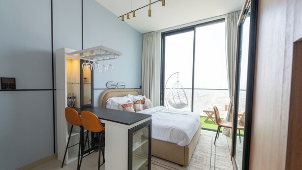 Ett modernt hotellrum med en säng, ett skrivbord med två stolar, ett litet kylskåp och ett stort fönster med utsikt över stranden.