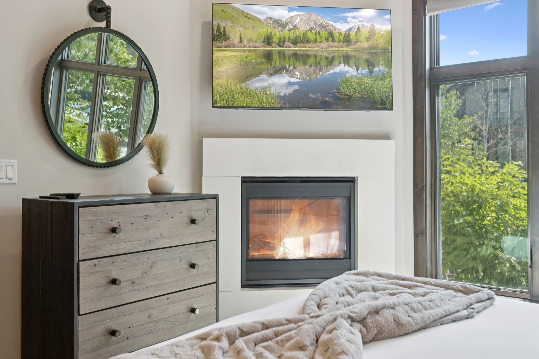 Ein Schlafzimmer mit Kamin, Kommode, Bett mit Decke, rundem Spiegel und großem Fenster mit Blick auf Bäume und Berge.
