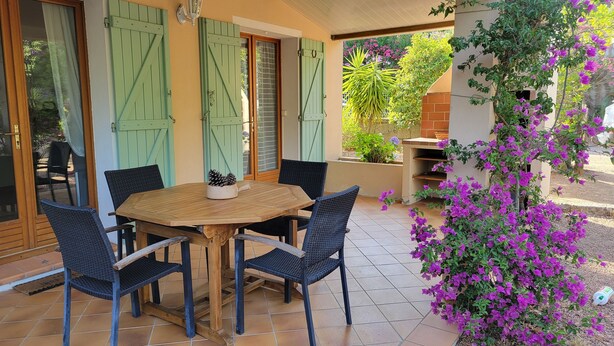 A covered patio with a wooden table and chairs, green shutters, and a potted plant with purple flowers. - South Corsica house, close to the most beautiful beaches and mountains (Sotta)