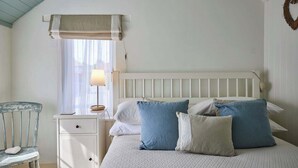 Un dormitorio con una cama blanca, una mesita de noche, una lámpara, una silla y una ventana con cortinas traslúcidas.