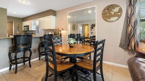 A dining area with a round table, black chairs, a kitchen in the background, and a mirror on the wall.