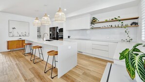 A modern kitchen with white cabinetry, a central island with stools, pendant lighting, and a large plant.