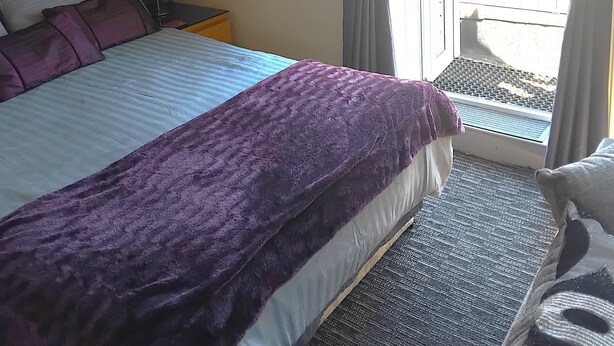 Habitación de hotel con cama, mesita de noche, lámpara, ventana con cortinas y vista al exterior.