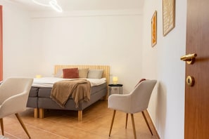 A bedroom with a bed, two chairs, a bedside table with a lamp, and a door with a golden handle.