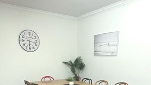 A dining area with a wooden table, six chairs, a clock, a potted plant, and a painting on the wall.