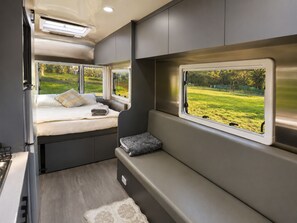 Un intérieur de camping-car compact comprenant un lit, une banquette et une fenêtre donnant sur un espace verdoyant.